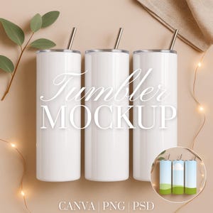 Op de afbeelding: Drie witte bekers met zilveren rietjes zijn gerangschikt op een beige oppervlak. De bekers hebben een cilindrische vorm en een glanzende afwerking. De tekst "Tumbler MOCKUP" wordt in cursieve letters weergegeven. De afbeelding bevat ook een takje met bladeren en lichtslingers.