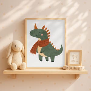 K&ouml;nnte beinhalten: Ein gerahmter Druck mit einem Cartoon-Dinosaurier mit einem rot-orangen Schal. Der Dinosaurier ist gr&uuml;n mit orangefarbenen Stacheln. Ein Stoffhase und ein kleiner gerahmter Schriftzug mit dem Text "DREAM BIG little one" befinden sich auf einem Regal.