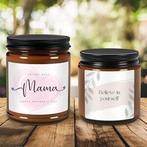 May include: Two brown glass candle jars with black lids. The jar on the left has a white label with a pink watercolour heart and the text "To the best Mama Happy Mother's Day". The jar on the right has a white label with a grey watercolour design and the text "Believe in yourself".