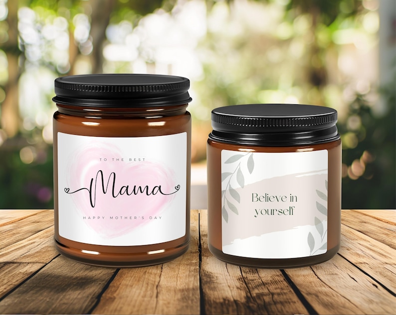 May include: Two brown glass candle jars with black lids. The jar on the left has a white label with a pink watercolour heart and the text "To the best Mama Happy Mother's Day". The jar on the right has a white label with a grey watercolour design and the text "Believe in yourself".