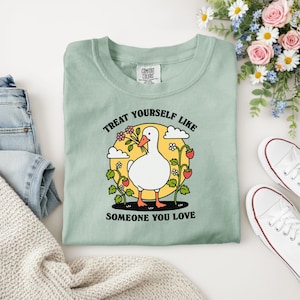 Puede incluir: Una camiseta verde claro Comfort Colors con un gr&aacute;fico de un ganso blanco sosteniendo flores, rodeado de fresas y el texto "TREAT YOURSELF LIKE SOMEONE YOU LOVE".