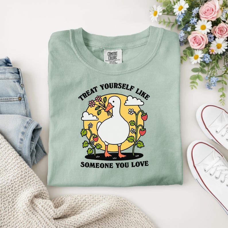 Puede incluir: Una camiseta verde claro Comfort Colors con un gr&aacute;fico de un ganso blanco sosteniendo flores, rodeado de fresas y el texto "TREAT YOURSELF LIKE SOMEONE YOU LOVE".