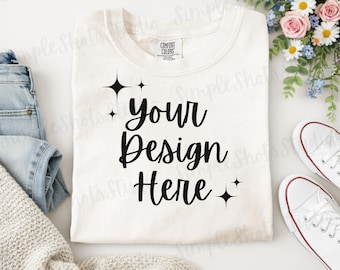 May include: Cream-coloured t-shirt with the text "Your Design Here" in black script. The shirt is folded and surrounded by white trainers, blue jeans, a grey sweater, and a bouquet of flowers. The shirt has a Comfort Colors tag.