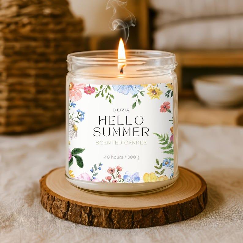 Op de afbeelding: Een brandende geurkaars in een glazen pot met een bloemenlabel met de tekst "HELLO SUMMER". De kaars staat op een houten onderzetter. Het label heeft een bloemenontwerp met roze, blauwe en gele bloemen. De kaars is ontworpen om 40 uur te branden en bevat 300 g was.
