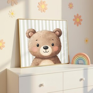 K&ouml;nnte beinhalten: Gerahmte Illustration eines Cartoon-B&auml;ren mit hellbraunem Fell, rosigen Wangen und einer schwarzen Nase. Der Hintergrund hat vertikale gr&uuml;ne und wei&szlig;e Streifen. Der Rahmen ist goldfarben. Das Kunstwerk wird auf einer wei&szlig;en Kommode mit einem Holzregenbogen ausgestellt.