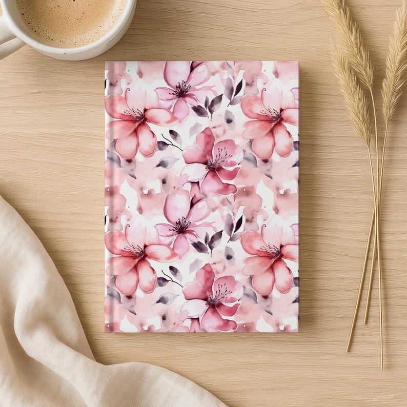 May include: A floral journal with a watercolor design of pink flowers and green leaves. The journal is rectangular and lies on a wooden surface, next to a cup of coffee and decorative wheat stalks.