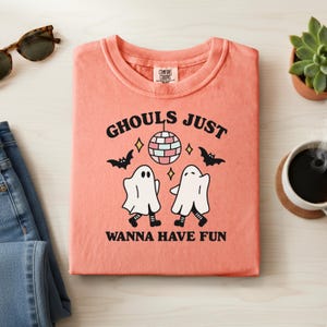 May include: Coral-colored sweatshirt with the text "Ghouls Just Wanna Have Fun" and a graphic of two dancing ghosts, a disco ball, and bats. The shirt is folded on a light wood surface, with sunglasses, jeans, and a succulent plant visible.