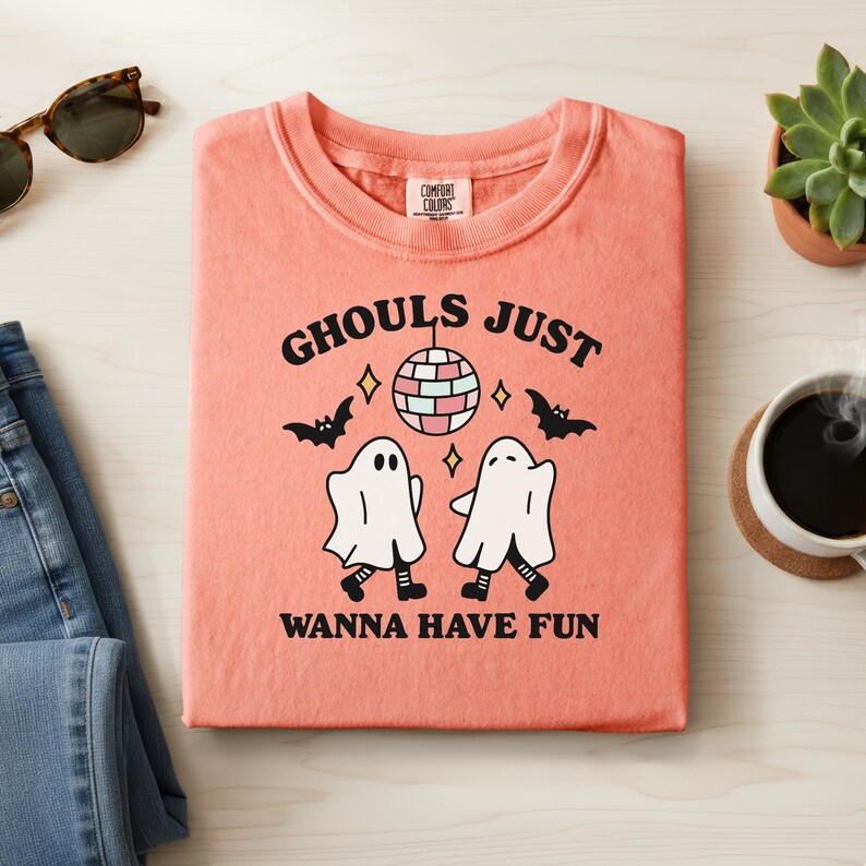 May include: Coral-colored sweatshirt with the text "Ghouls Just Wanna Have Fun" and a graphic of two dancing ghosts, a disco ball, and bats. The shirt is folded on a light wood surface, with sunglasses, jeans, and a succulent plant visible.