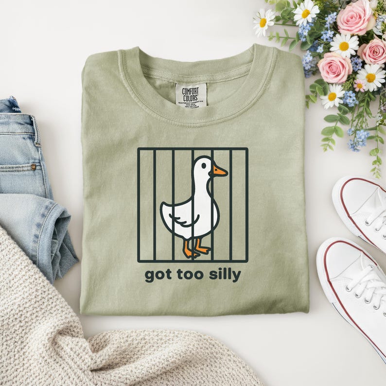 Puede incluir: Camiseta verde salvia con un dibujo de un pato de dibujos animados tras las rejas, con el texto "got too silly" debajo. La camiseta est&aacute; junto a unos vaqueros azules, un su&eacute;ter crema y zapatillas blancas.
