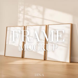 May include: Three rectangular picture frames with a light wood finish are leaning against a beige wall. The frames have white interiors and the words "FRAME MOCKUP" are displayed in large white letters across the frames. The floor is a light wood colour.