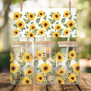 May include: Three clear glass jars with wooden lids and straws, decorated with a sunflower pattern. The jars are on a wooden surface with a blurred background of green foliage.