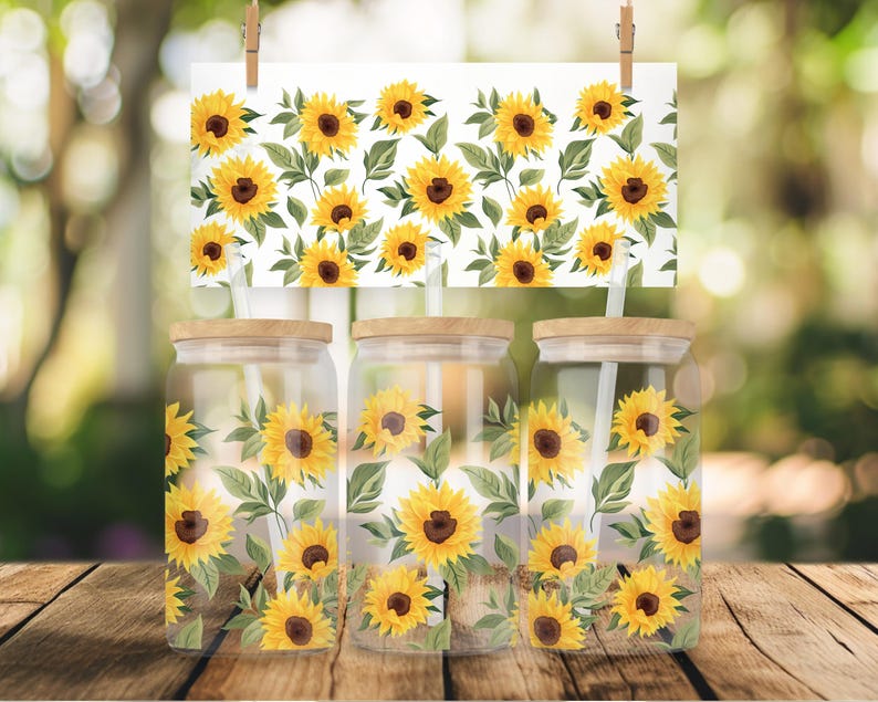 May include: Three clear glass jars with wooden lids and straws, decorated with a sunflower pattern. The jars are on a wooden surface with a blurred background of green foliage.