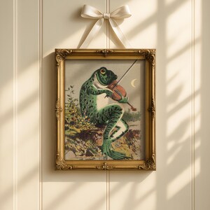 Op de afbeelding: Een ingelijste vintage prent toont een kikker die viool speelt. De kikker, met een groen-zwarte gespikkelde huid, zit op een rots. De prent zit in een gouden, sierlijke lijst, opgehangen aan een cr&egrave;mekleurig lint met een strik.