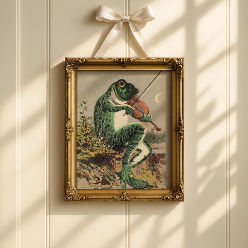 Op de afbeelding: Een ingelijste vintage prent toont een kikker die viool speelt. De kikker, met een groen-zwarte gespikkelde huid, zit op een rots. De prent zit in een gouden, sierlijke lijst, opgehangen aan een cr&egrave;mekleurig lint met een strik.
