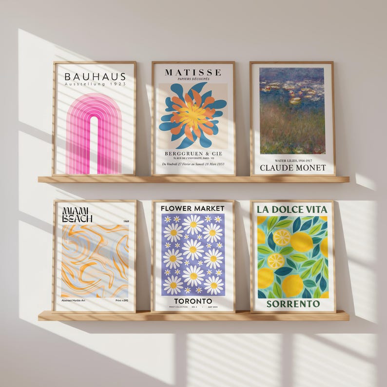 May include: Six framed art prints displayed on wooden shelves. The prints feature various designs, including a pink arch, a floral abstract, a Monet painting, a Miami Beach design, a daisy pattern, and a lemon illustration. The frames are a light brown color.
