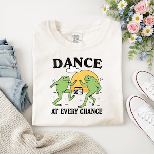 Puede incluir: Camiseta color crema con la frase "DANCE AT EVERY CHANCE" en negro. La camiseta presenta un gr&aacute;fico de dos ranas verdes bailando frente a un sol amarillo y una radio.