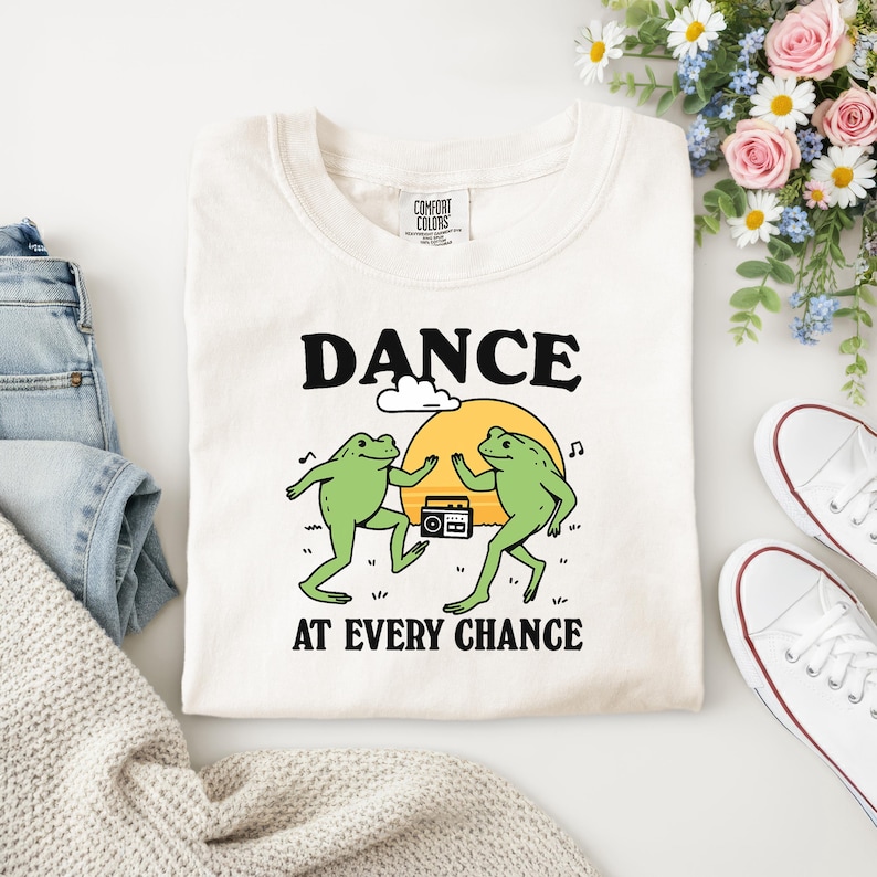 Puede incluir: Camiseta color crema con la frase "DANCE AT EVERY CHANCE" en negro. La camiseta presenta un gr&aacute;fico de dos ranas verdes bailando frente a un sol amarillo y una radio.
