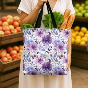 May include: A reusable tote bag with a floral pattern featuring purple and blue flowers on a white background. The bag is being held by a person in a grocery store.