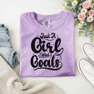 Puede incluir: Una camiseta de color lila con el texto "Just A Girl With Goals" en letras negras. La camiseta est&aacute; doblada sobre una superficie blanca, junto a zapatillas blancas y un ramo de flores. La camiseta tiene una etiqueta Comfort Colors.