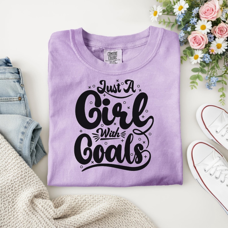 Puede incluir: Una camiseta de color lila con el texto "Just A Girl With Goals" en letras negras. La camiseta est&aacute; doblada sobre una superficie blanca, junto a zapatillas blancas y un ramo de flores. La camiseta tiene una etiqueta Comfort Colors.