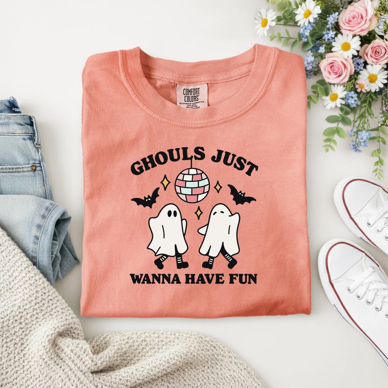 Puede incluir: Camiseta Comfort Colors de color coral con el texto "Ghouls Just Wanna Have Fun". El dise&ntilde;o incluye dos figuras de fantasmas, una bola de discoteca, murci&eacute;lagos y diamantes. La camiseta se muestra con vaqueros y un jersey.