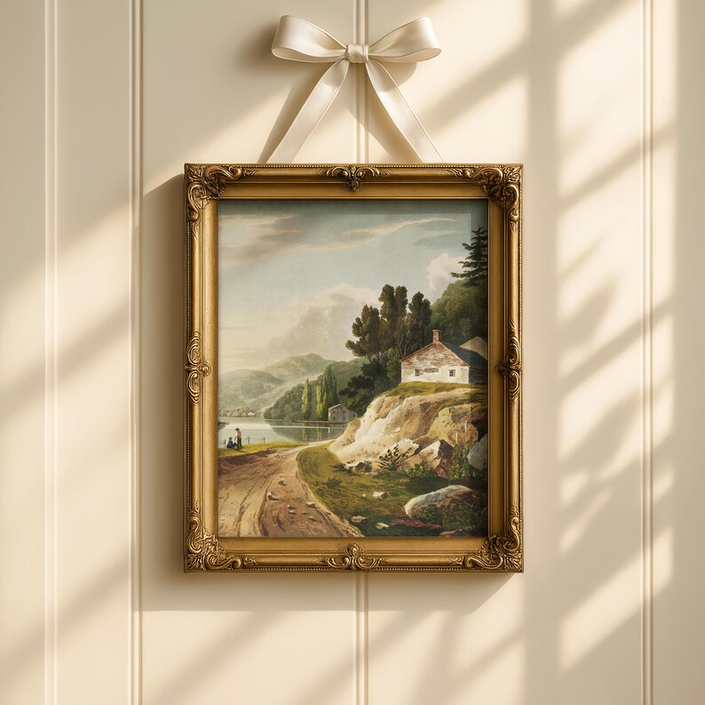Op de afbeelding: Een ingelijst landschapsschilderij met een gouden, sierlijke lijst. Het kunstwerk toont een serene sc&egrave;ne met een onverharde weg, een klein wit huis en een meer omgeven door heuvels. Bovenop is een cr&egrave;mekleurig lint geknoopt om op te hangen.