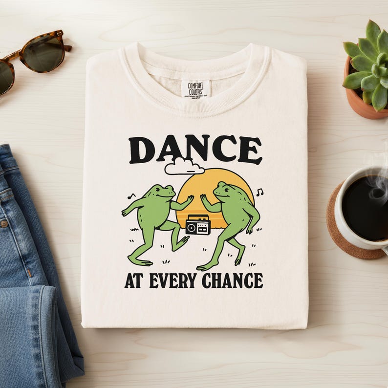 May include: Cream-colored sweatshirt with a graphic of two green frogs dancing in front of a yellow sun. The text "DANCE AT EVERY CHANCE" is above and below the frogs. The sweatshirt is folded on a light-colored surface.