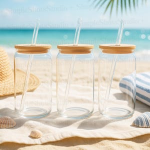 Könnte beinhalten: Drei klare Glasbecher mit Holzdeckeln und klaren Trinkhalmen. Die Becher stehen auf einem weißen Strandtuch auf einem Sandstrand.