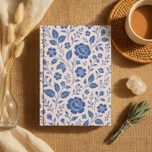May include: A hardcover journal with a cream-colored background and a blue floral pattern. The design features detailed flowers, leaves, and vines. The journal is on a textured surface with a cup of coffee, a crystal, and a bundle of sage.