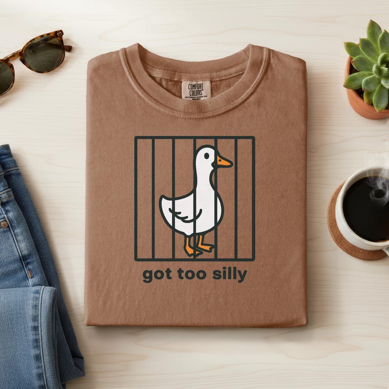 May include: A brown t-shirt with a crew neck featuring a cartoon duck behind bars. The duck is white with an orange beak and feet. The text "got too silly" is below the graphic. The shirt is folded on a light wood surface.