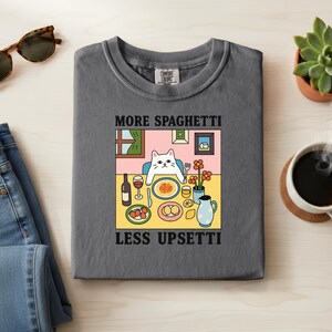 May include: A gray Comfort Colors t-shirt featuring a whimsical illustration of a cat at a table set for a meal. The text "MORE SPAGHETTI LESS UPSETTI" is printed above and below the illustration. The cat is sitting at a table with food and drinks.