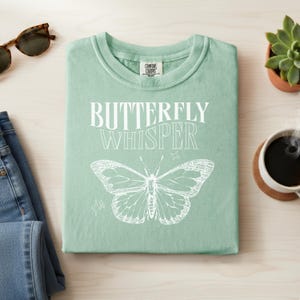 May include: A light green t-shirt with the words "BUTTERFLY WHISPER" in white, along with a detailed butterfly illustration. The shirt is folded neatly on a light wood surface, with sunglasses and jeans nearby.