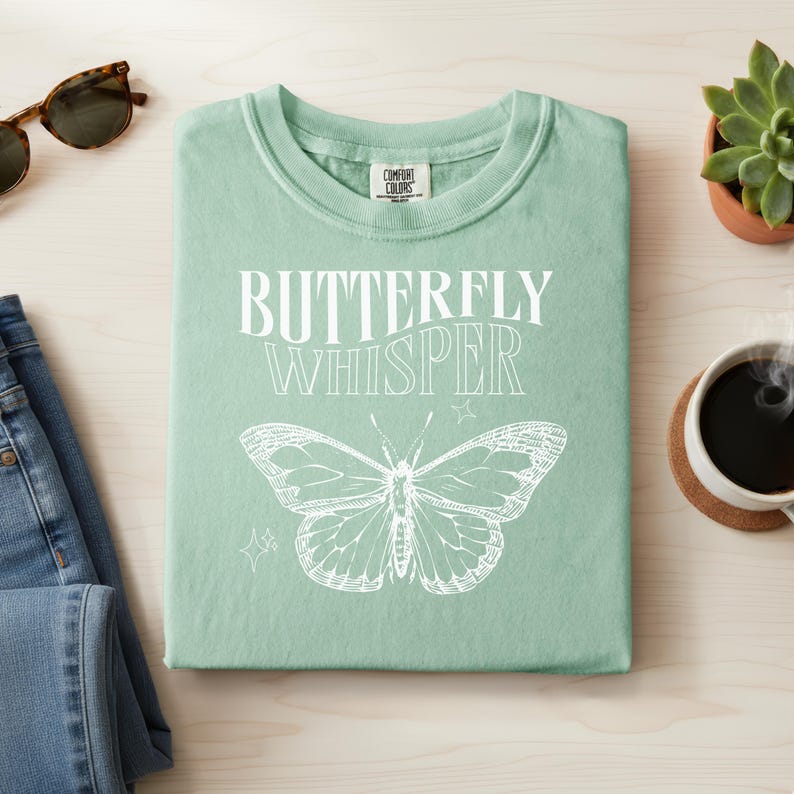May include: A light green t-shirt with the words "BUTTERFLY WHISPER" in white, along with a detailed butterfly illustration. The shirt is folded neatly on a light wood surface, with sunglasses and jeans nearby.