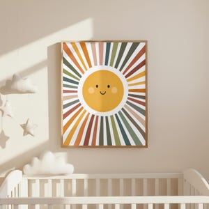 K&ouml;nnte beinhalten: Ein gerahmter Kunstdruck mit einer l&auml;chelnden Sonne mit farbenfrohen Strahlen in Orange-, Gr&uuml;n- und Braunt&ouml;nen. Die Sonne hat ein gelbes Gesicht mit einem gebogenen Mund und kleinen schwarzen Punkten f&uuml;r die Augen. Das Kunstwerk wird &uuml;ber einem wei&szlig;en Kinderbett gezeigt.