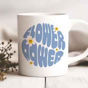 K&ouml;nnte beinhalten: Wei&szlig;e Keramiktasse mit einem blauen und wei&szlig;en Design, auf dem in einer Retro-Schrift "Flower Power" steht. Das Design ist von kleinen wei&szlig;en G&auml;nsebl&uuml;mchen mit gelben Smiley-Gesichtern umgeben.