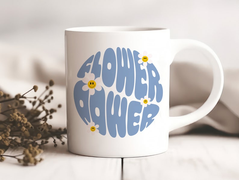 K&ouml;nnte beinhalten: Wei&szlig;e Keramiktasse mit einem blauen und wei&szlig;en Design, auf dem in einer Retro-Schrift "Flower Power" steht. Das Design ist von kleinen wei&szlig;en G&auml;nsebl&uuml;mchen mit gelben Smiley-Gesichtern umgeben.