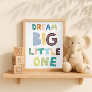 K&ouml;nnte beinhalten: Ein gerahmter Druck mit den Worten "DREAM BIG LITTLE ONE" in bunten Buchstaben steht auf einem Holzregal. Daneben ein Stoffelefant und ABC-Bl&ouml;cke aus Holz. Ein kleines Holzhaus steht ebenfalls auf dem Regal. Szene f&uuml;r ein Kinderzimmer.