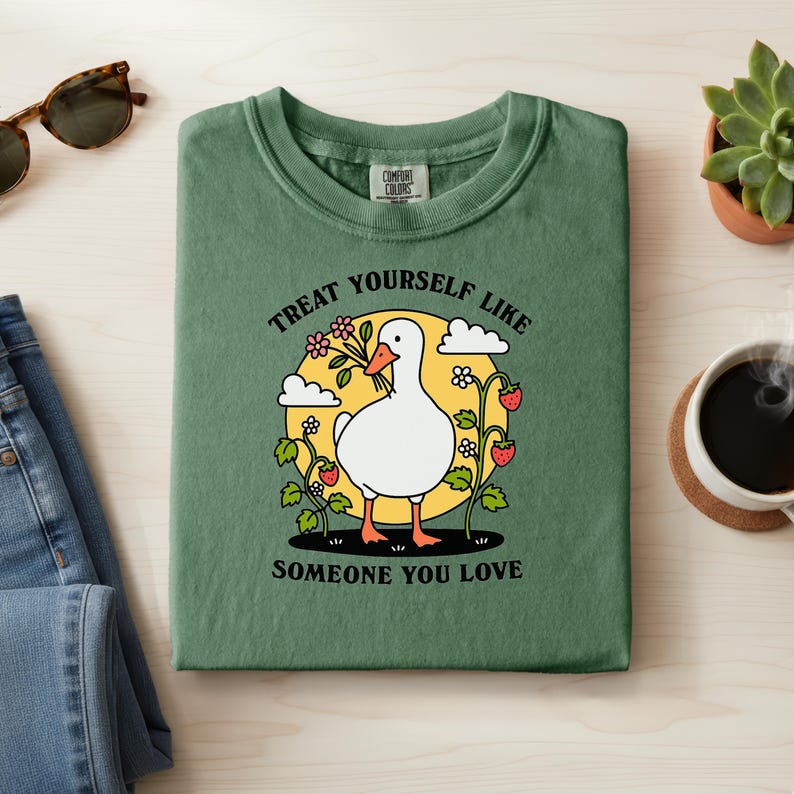 May include: Green t-shirt with a graphic of a white goose holding flowers, surrounded by strawberries and the text "Treat Yourself Like Someone You Love". The shirt is folded on a light surface.