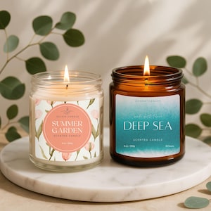 May include: Two lit scented candles in glass jars. One is labeled "Summer Garden" with a floral design, the other "Deep Sea" with a blue watercolor effect. Both are on a white marble surface.