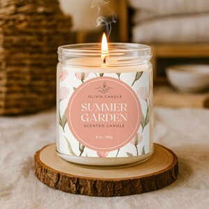 Op de afbeelding: Een brandende geurkaars in een glazen pot met een bloemenlabel met de tekst "Summer Garden". De kaars staat op een houten onderzetter. Het label bevat de tekst "Olivia Candle" en de kaars bevat 250g was.