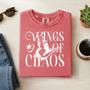 May include: A coral-colored t-shirt with the words "WINGS OF CHAOS" and a butterfly graphic in white. The shirt is folded neatly on a light wood surface, with sunglasses, jeans, a succulent, and a cup of coffee nearby.