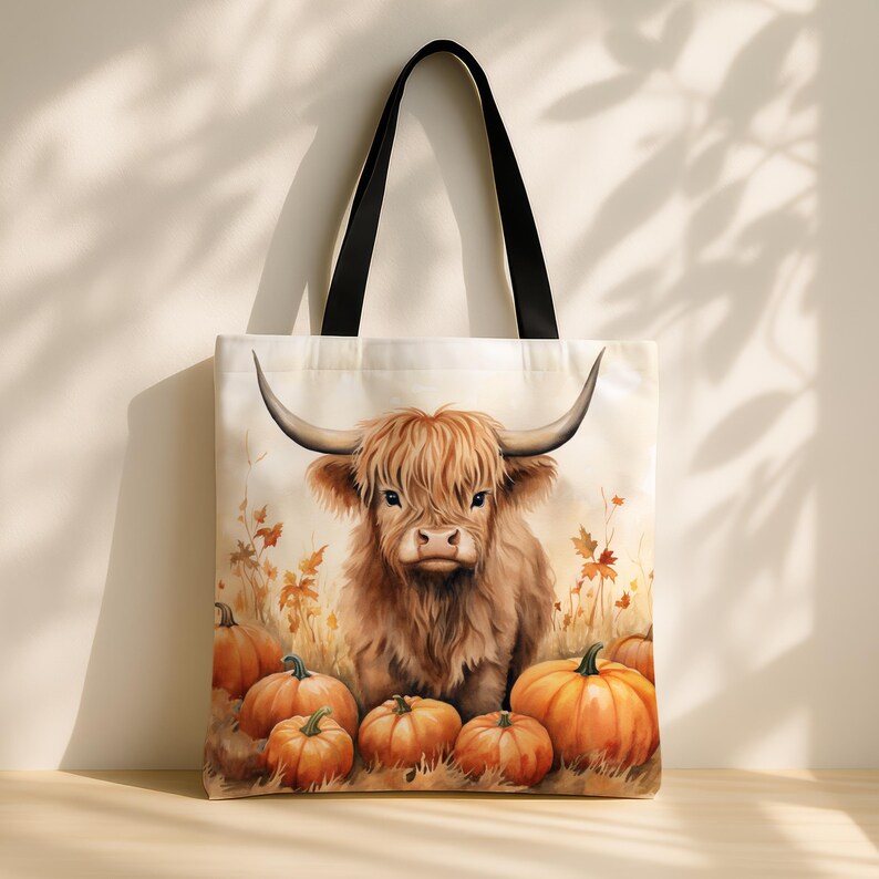 Puede incluir: Una bolsa de lona blanca con una ilustraci&oacute;n de una vaca Highland marr&oacute;n y blanca y un dise&ntilde;o de calabazas naranjas. La bolsa tiene asas negras.