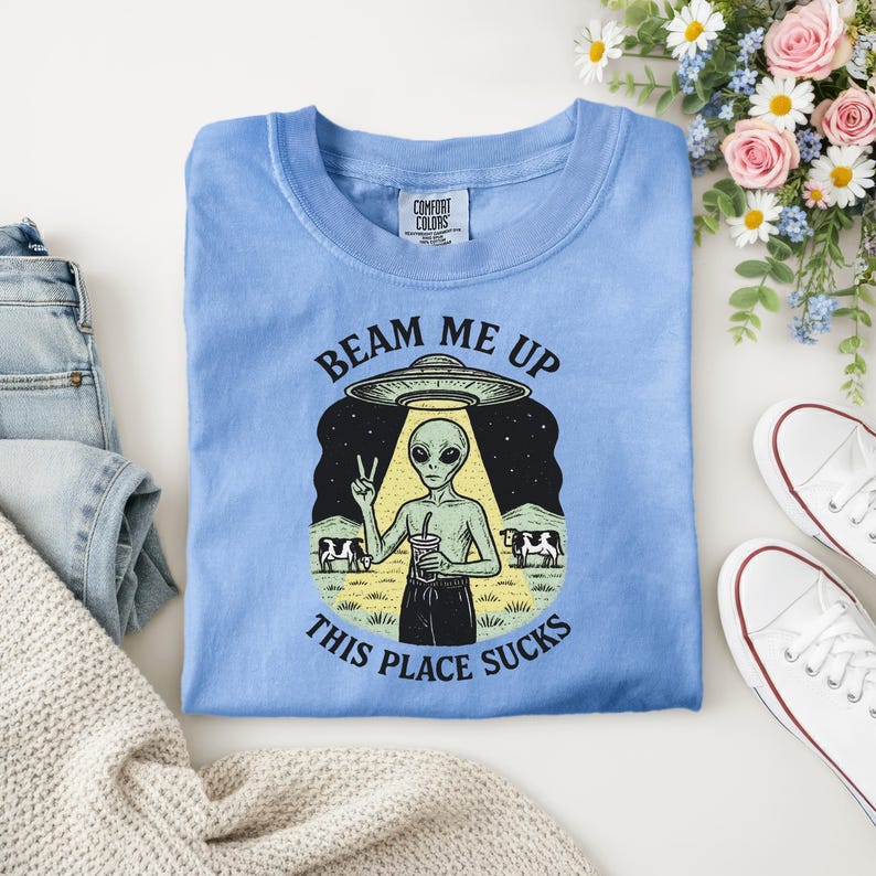 Puede incluir: Camiseta azul claro con un gr&aacute;fico de un extraterrestre bajo un OVNI, sosteniendo una bebida y haciendo el signo de la paz. El texto dice "Beam Me Up, This Place Sucks". Hecho de material Comfort Colors.