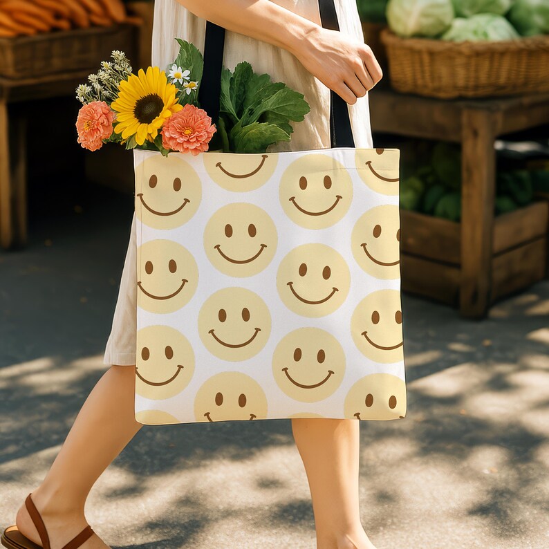 Puede incluir: Una bolsa de tela blanca con un patr&oacute;n repetido de caras sonrientes amarillas. La bolsa la lleva una persona que lleva un vestido beige y un ramo de flores.