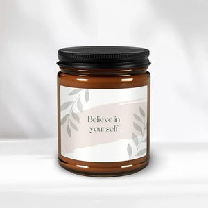 May include: A brown glass candle jar with a black lid and a white label. The label has a light pink watercolour background with grey leaves and the text "Believe in yourself".