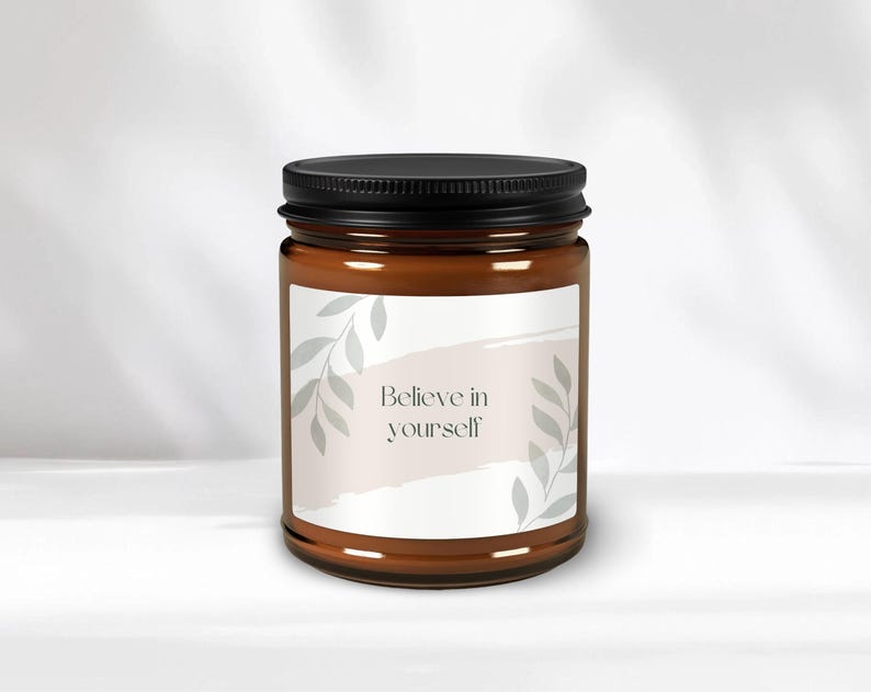 May include: A brown glass candle jar with a black lid and a white label. The label has a light pink watercolour background with grey leaves and the text "Believe in yourself".