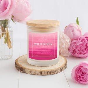 Może przedstawiać: Świeca zapachowa Wild Berry z drewnianą pokrywką, prezentowana na drewnianym plastrze. Etykieta zawiera akwarelowy wz&oacute;r w odcieniach r&oacute;żu i napis "Wild Berry". Świeca otoczona jest r&oacute;żowymi piwoniami.