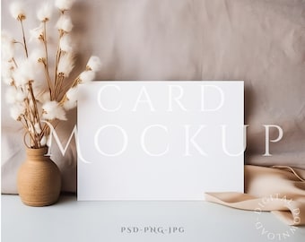 May include: A white card mockup with the text "CARD MOCKUP" in a white sans-serif font. The card is sitting on a light blue surface with a beige fabric background. A vase with dried white cotton flowers is on the left side of the image.