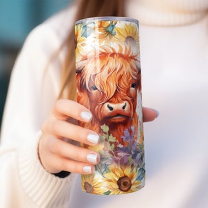 May include: A stainless steel tumbler with a colorful design featuring a Highland cow with brown fur and a white face surrounded by sunflowers and other flowers.
