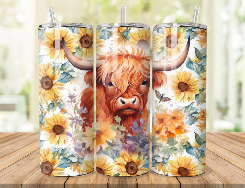 Puede incluir: Tres vasos de acero inoxidable con un dise&ntilde;o de una vaca Highland de pelaje marr&oacute;n y cara blanca, rodeados de girasoles y otras flores. Los vasos est&aacute;n sobre una superficie de madera.